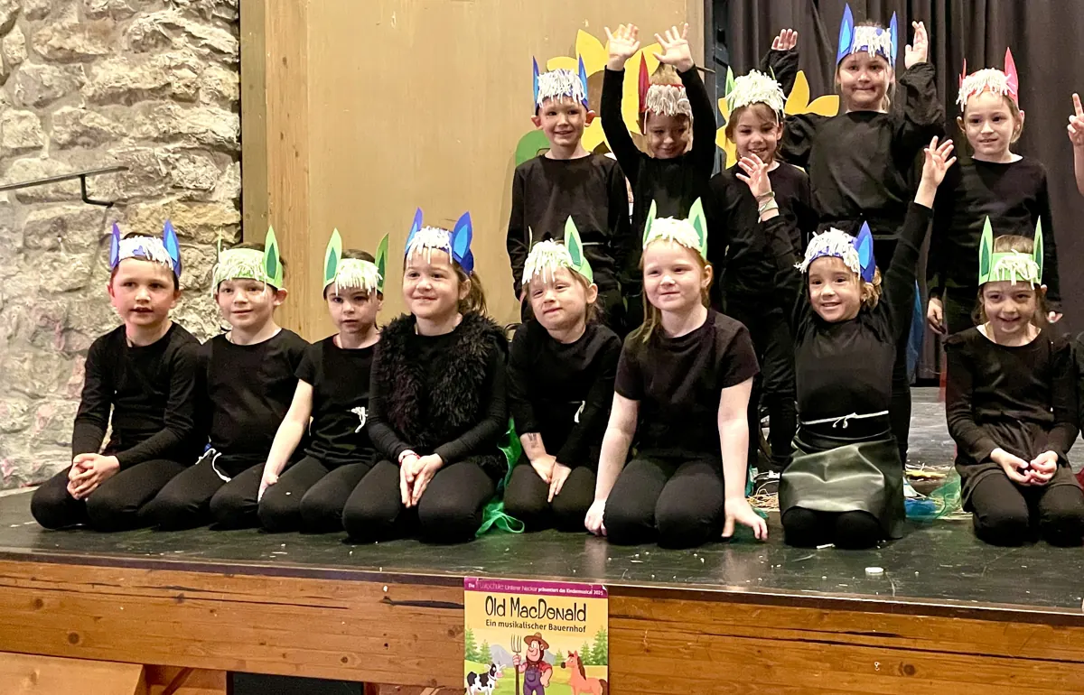 Kinder der Musikalischen Früherziehung in Schwarz und mit gebastelten Tierohren auf der Bühne, vorn das Plakat zum Kindermusical „Old MacDonald“