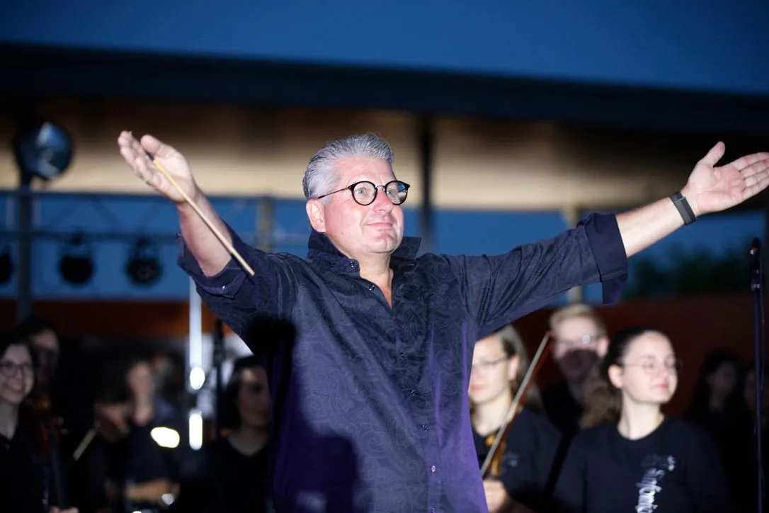 Schulleiter Marco Rogalski breitet mit dem Taktstock in der Hand die Arme aus, hinter ihm junge Musikerinnen und Musiker des Orchesters
