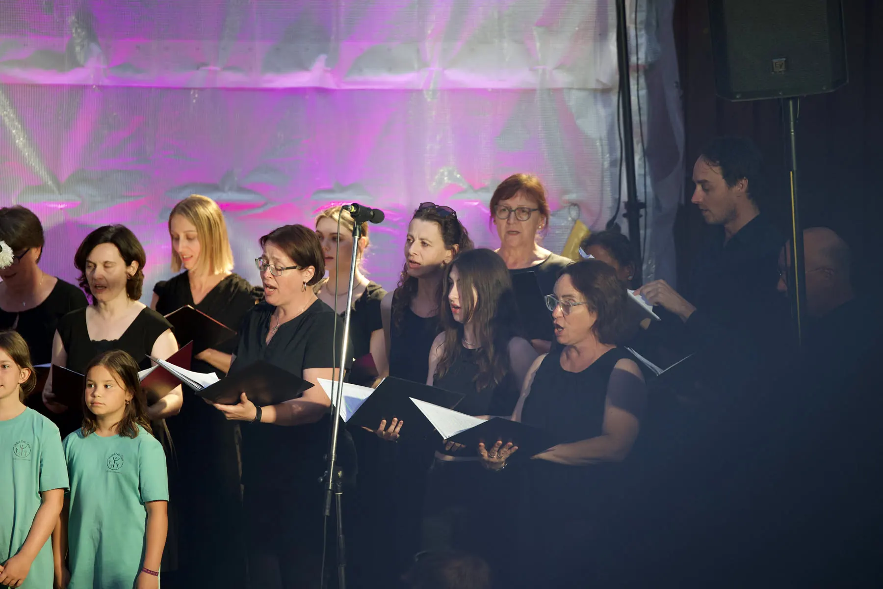 Sängerinnen und Sänger des Projektchors singen mit Notenmappen auf der Bühne, vorn zwei Kinder aus dem Kinderchor