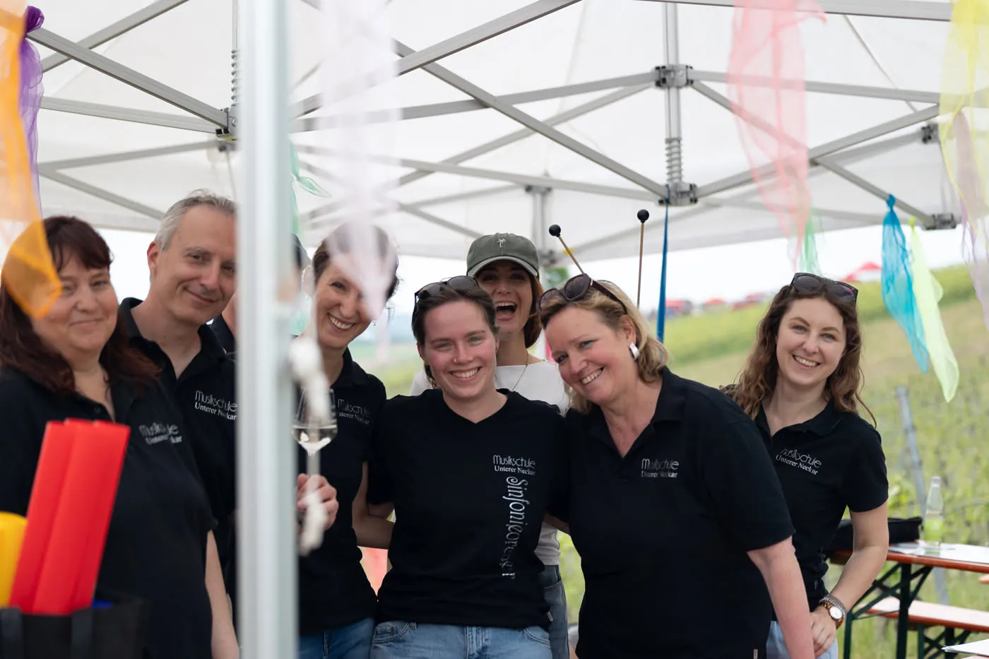 Sieben Kolleginnen und Kollegen in schwarzen Shirts der Musikschule lachen unter einem Zelt mit bunten Tüchern